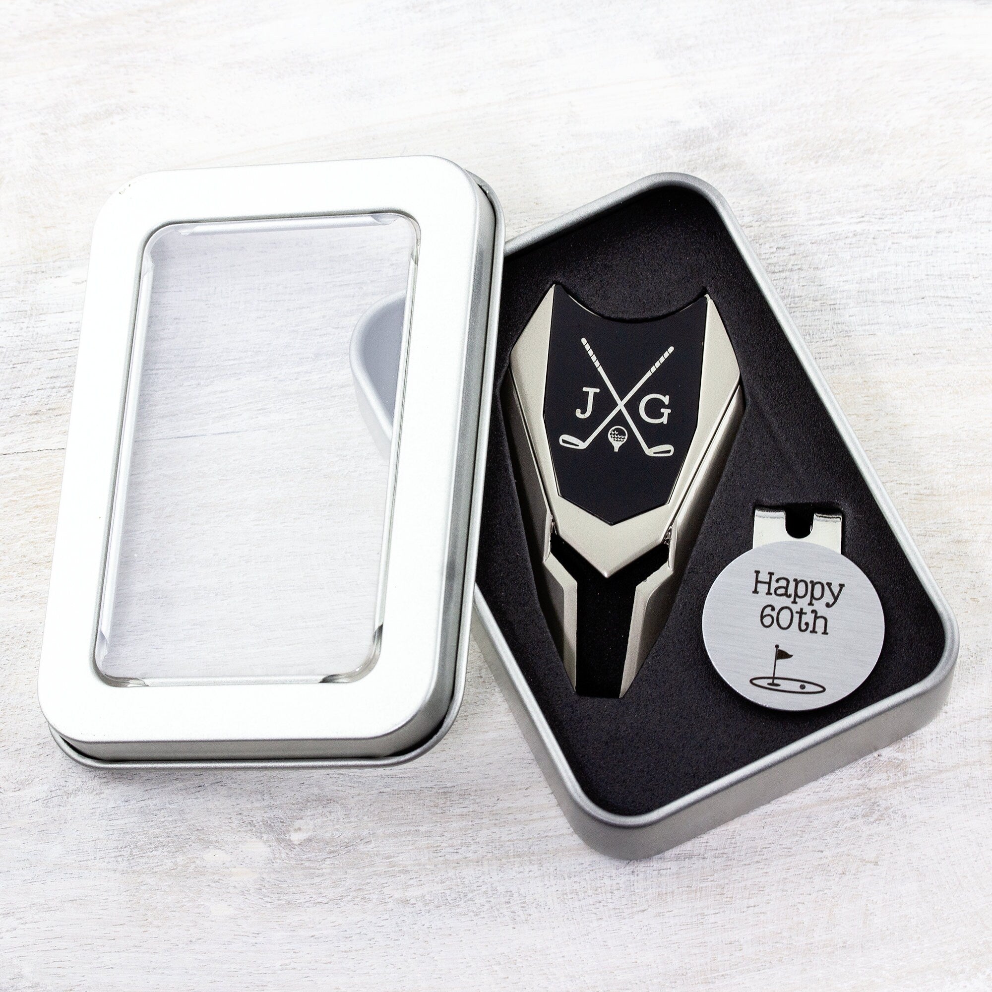Golf ball marker and divot tool gift tin set personalized. Divot tool has crossed clubs with initials on each side. Round ball marker with hat clip has happy 60th with golf flag graphic engraved.