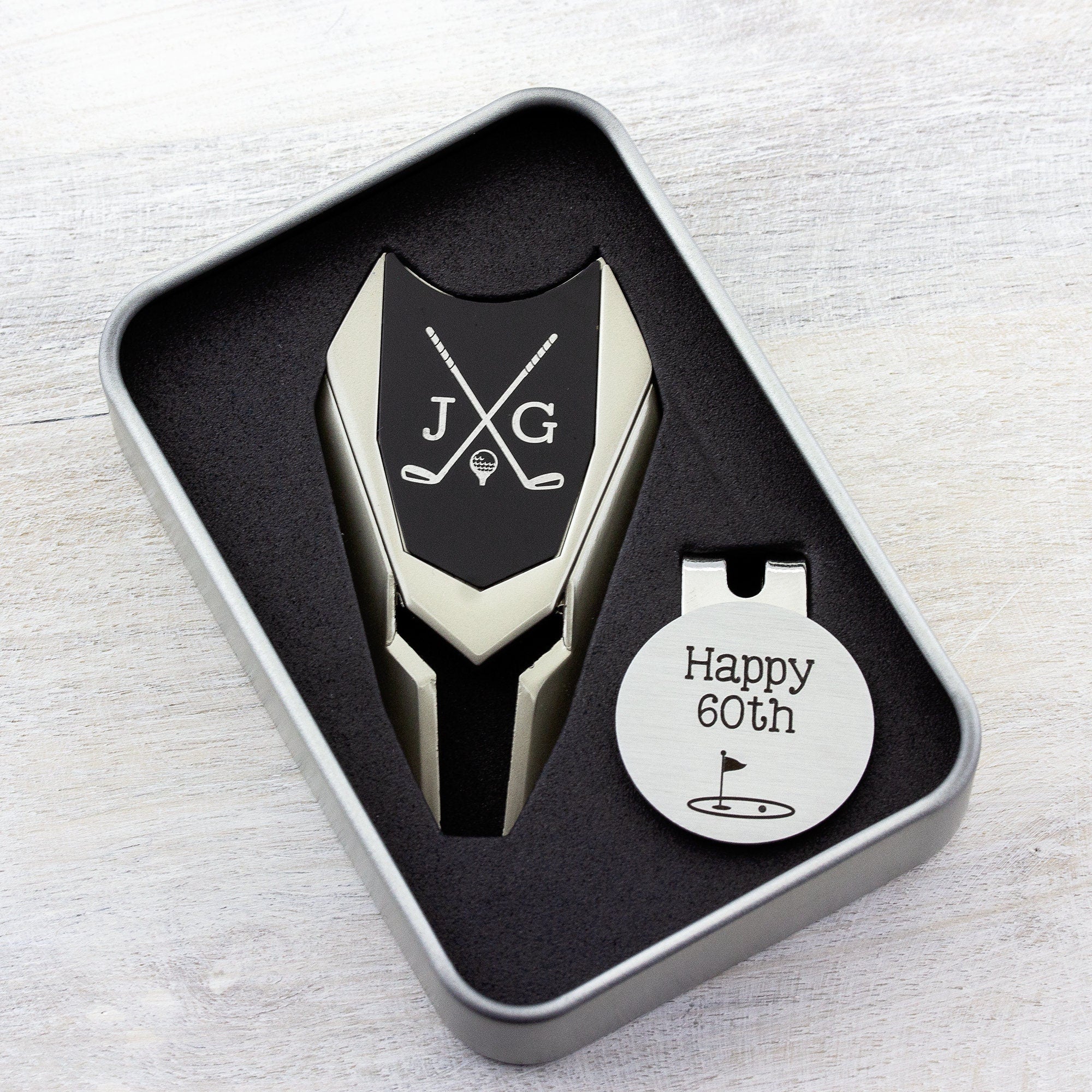Golf ball marker and divot tool gift tin set personalized. Divot tool has crossed clubs with initials on each side. Round ball marker with hat clip has happy 60th with golf flag graphic engraved.